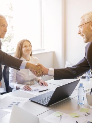 smiling-woman-looking-two-senior-businessman-shaking-hands